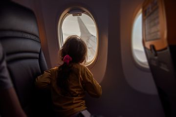 Little girl looks out window of airplane at wing. Child is not afraid to fly on plane, looks at sky through porthole. Curiosity, courage of children when flying. Atmosphere of airplane from the inside