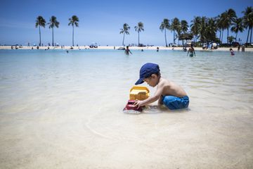 A two year old child plays with a toy in the water on the beach in Hawaii, wearing blue swim shorts and blue baseball cap.