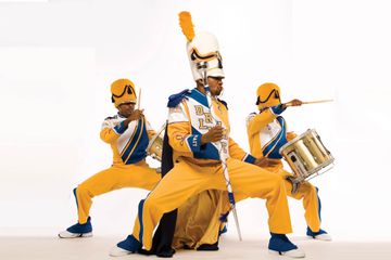 Black drumline group, the leader and two drummers in yellow and blue uniforms on white backdrop