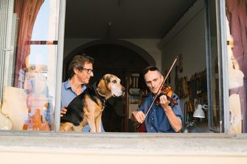 A violin maker and his dog watch a violinist playing in a studio window