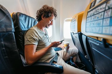 Handsome young man using smart phone and drinking coffee while in an Airplane.