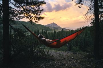A person laying in a red hammock between two tall pine trees with a mountain in the distance and birds flying