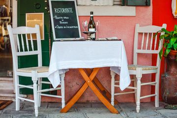Italian ristorante restaurant served table with white table-cloth and wooden chairs. Old vintage furniture. Wine goblets. Cafe serving for supper in Italy on the street