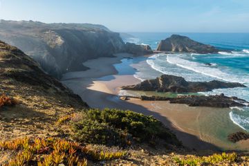 View of empty Praia da Zambujeira do Mar beach with ocean waves, cliffs and stones, wet golden sand and green vegetation at wild Rota Vicentina coast, Odemira, Portugal. 